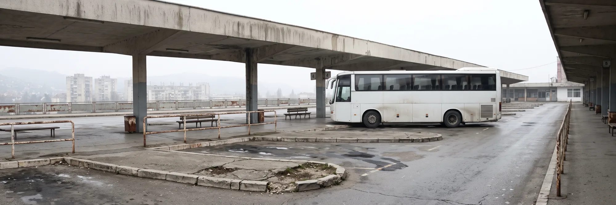 Një autobus i bardhë i parkuar në një peron të zbrazët nën strehën e gjatë prej betoni të një stacioni autobusësh, me bankina, parmakë të ndryshkur dhe pallate në mjegull në sfond, në një mëngjes me re