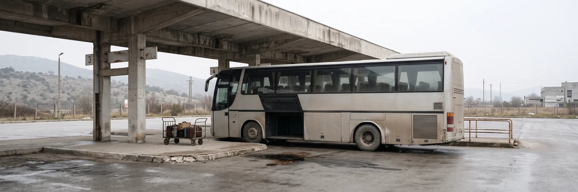 Një autobus i gjatë linje i parkuar nën strehën prej betoni të një stacioni autobusësh, me derën e bagazheve të hapur, një karrocë me valixhe pranë platformës dhe kodra të mjegulluara në sfond, në një ditë me re pas shiut
